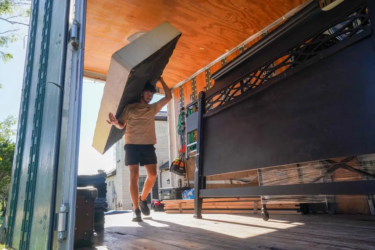 Trinity Movers crew member carrying a padded furniture piece overhead past a large bed frame secured inside a moving truck