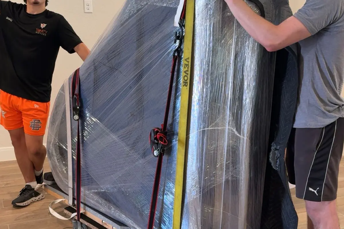 Two Trinity Movers crew members steadying a tall shrink-wrapped piece of furniture strapped to a dolly inside a home