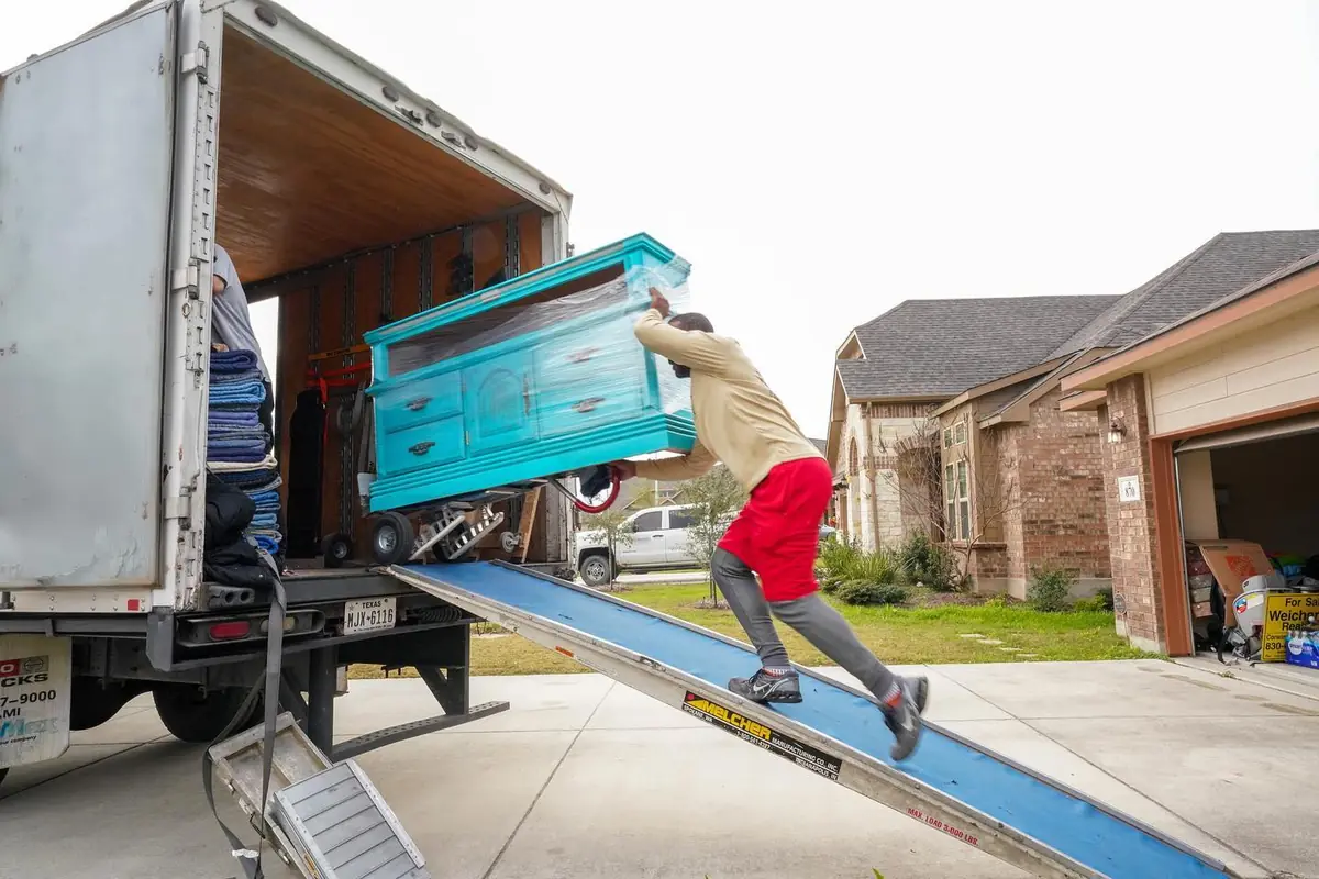 Trinity Movers crew member pushing a turquoise dresser up a loading ramp into a truck in a residential driveway