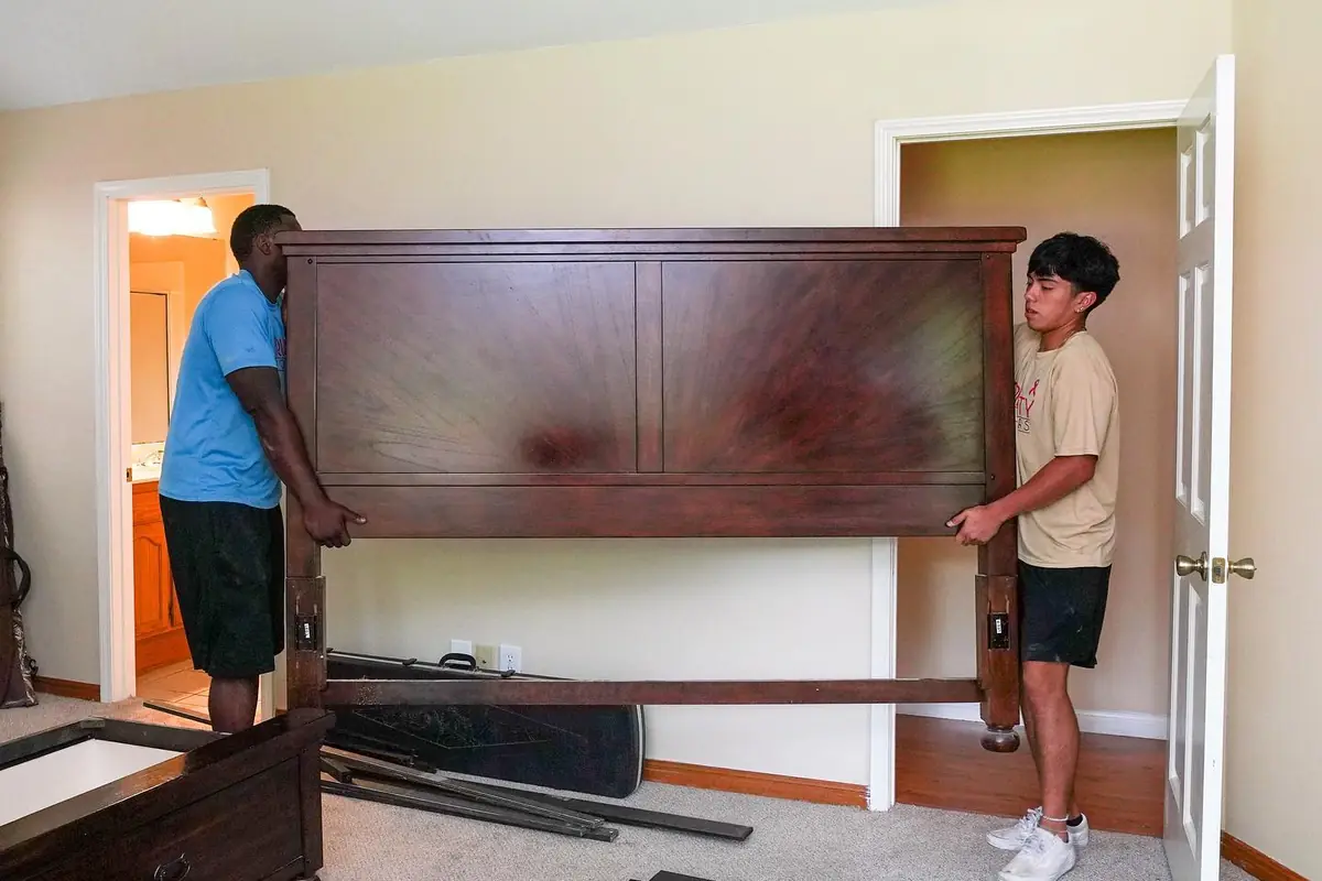 Two Trinity Movers crew members carrying a wooden headboard through a bedroom beside disassembled bed rails