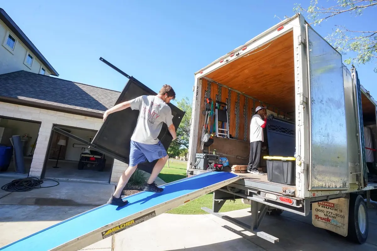 Trinity Movers crew member carrying a dark headboard up a ramp into an open box truck in a residential driveway