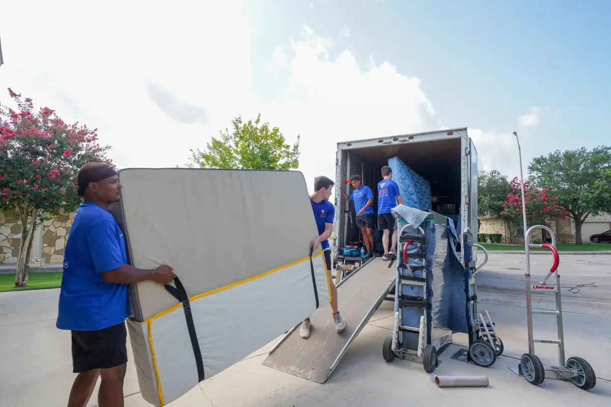 Trinity Movers crew member steadying a strapped mattress beside a loading ramp and an open moving truck