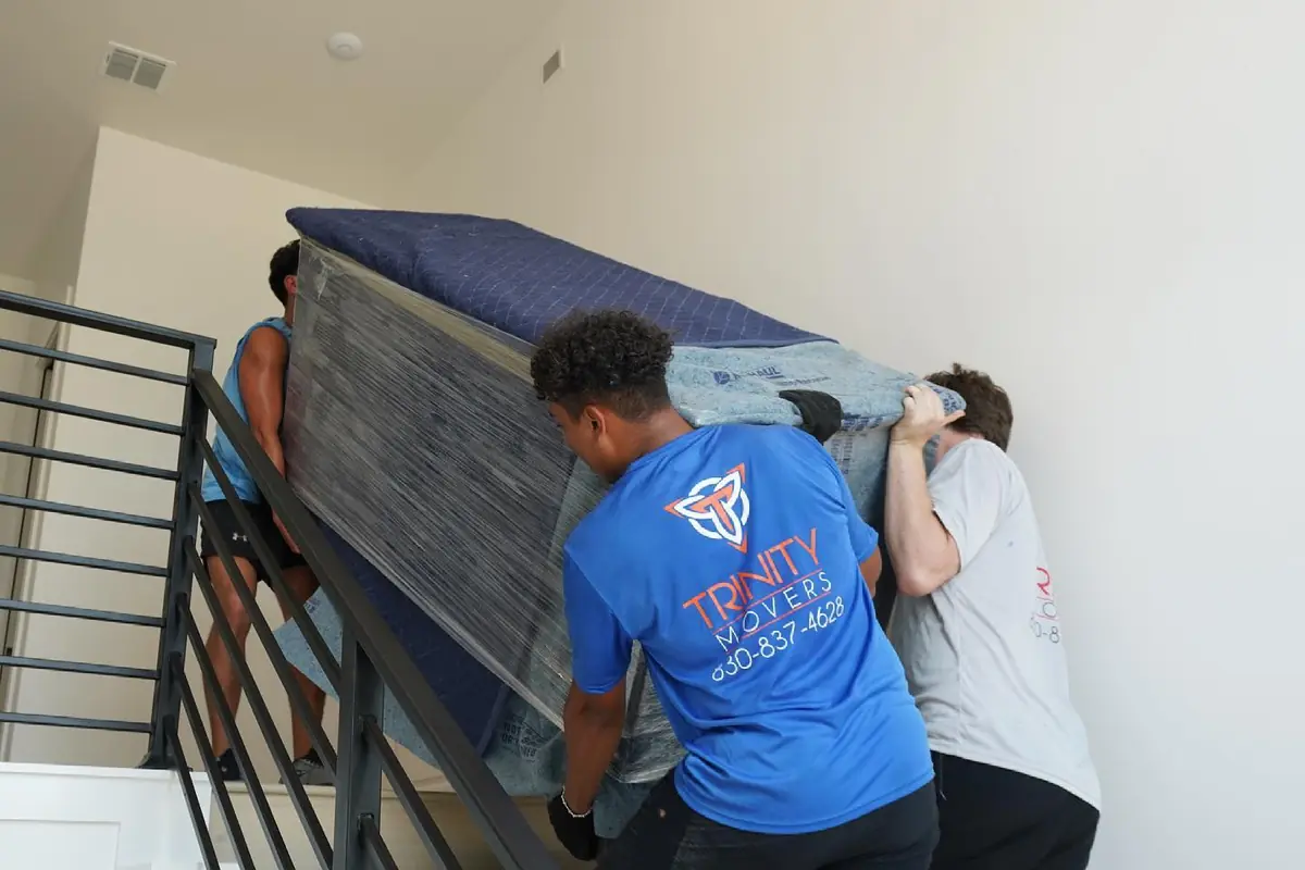 Three Trinity Movers crew members carrying a blanket-wrapped piece of furniture up an interior staircase