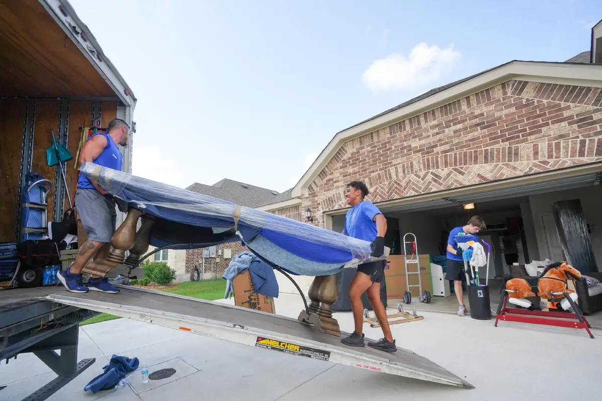 Two Trinity Movers crew members carrying a blanket-wrapped dining table down a truck ramp in front of a brick home