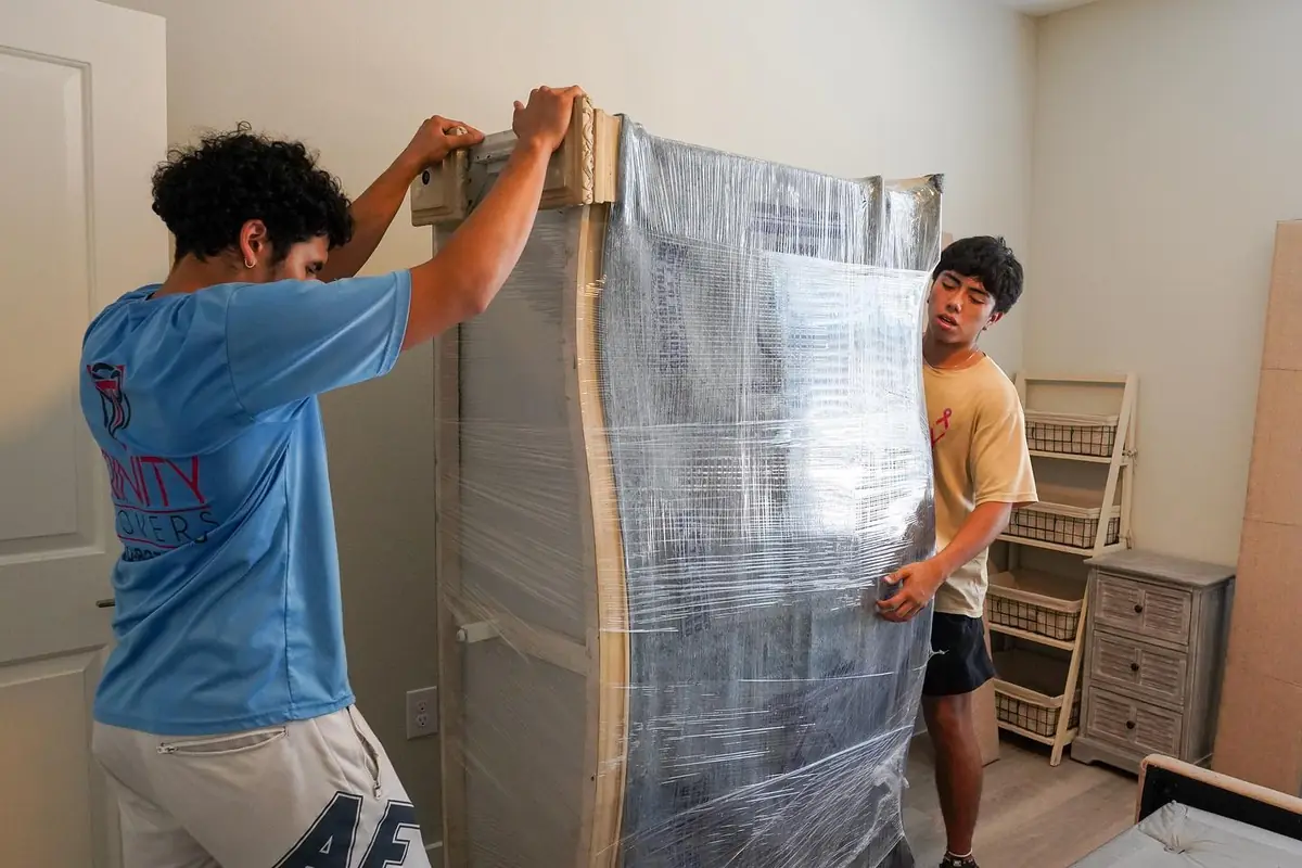 Two Trinity Movers crew members stretch-wrapping a tall padded piece of furniture in a bedroom