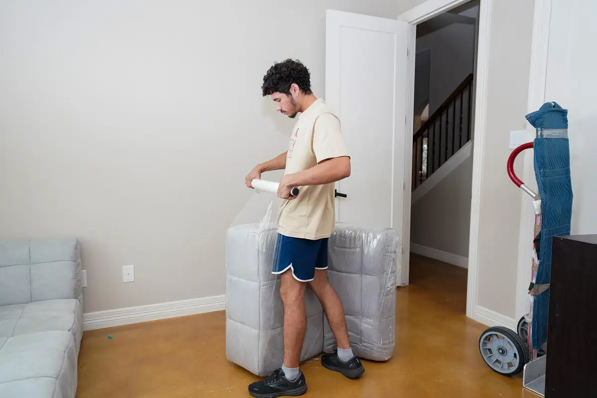 Trinity Movers crew member wrapping stacked cushions in stretch wrap in a living room beside a hand truck